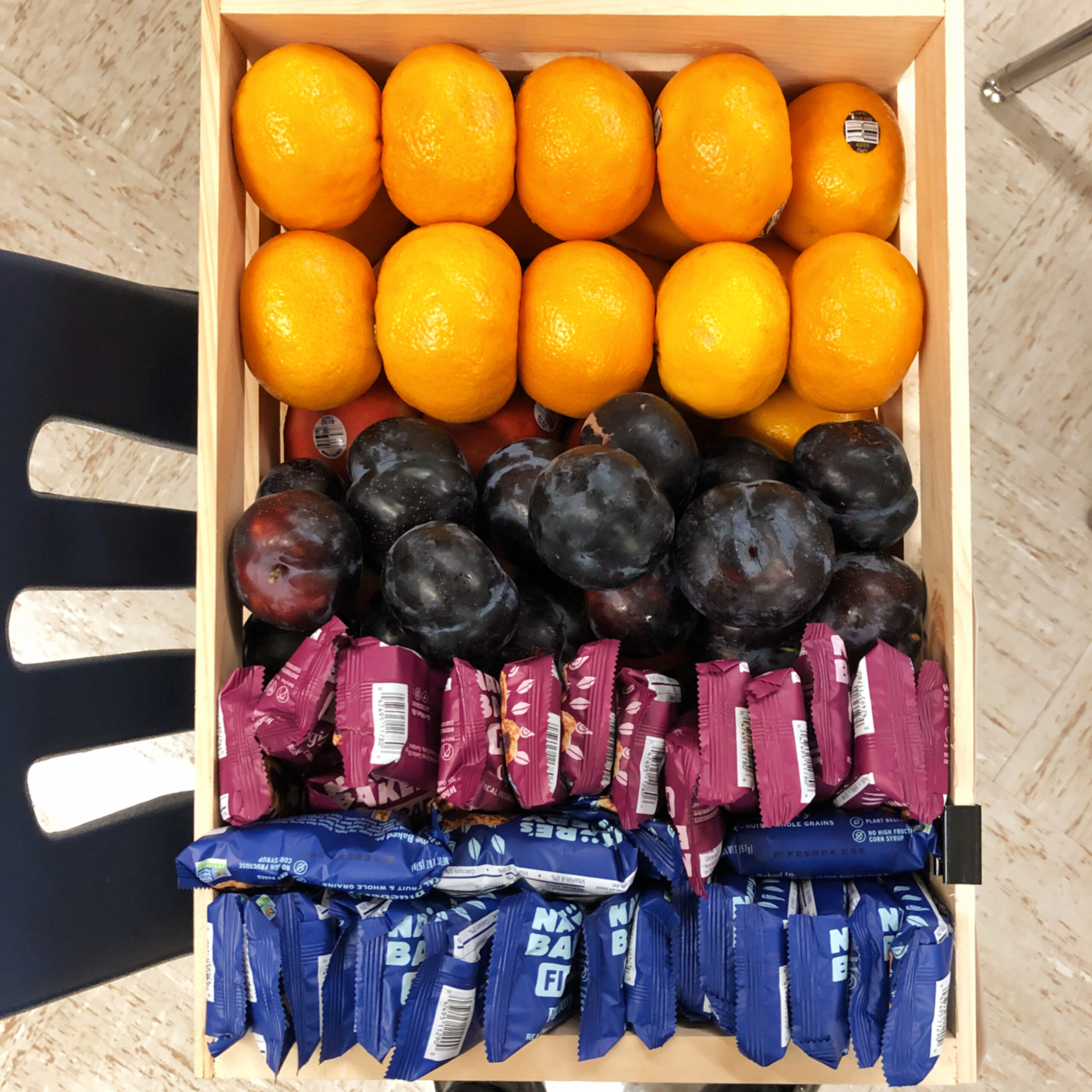 image is of a box of healthy snacks: oranges, plums, and fig bars. 